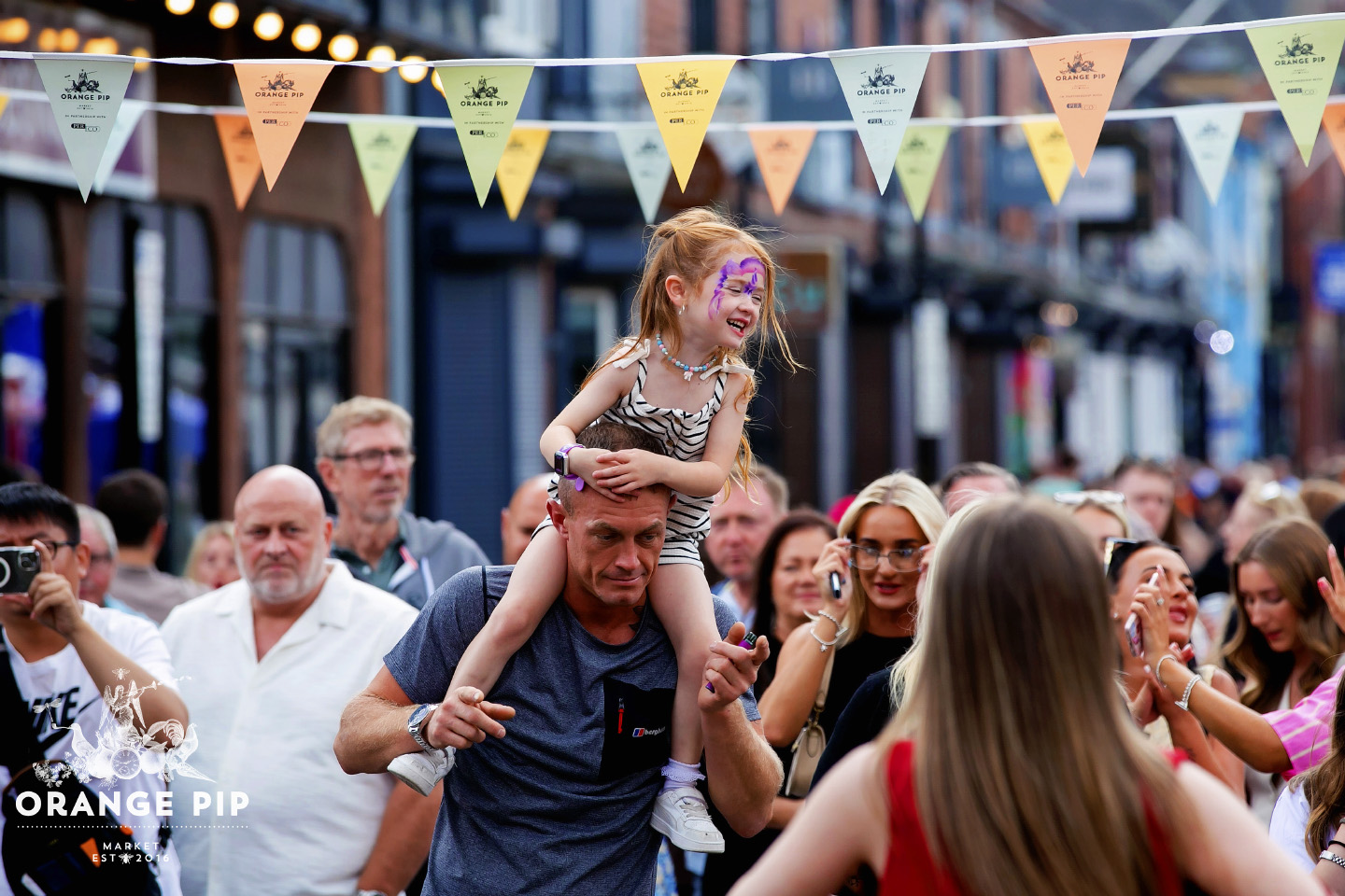 A child on the shoulders of an adult in a crowded orange pip market with bunting overhead