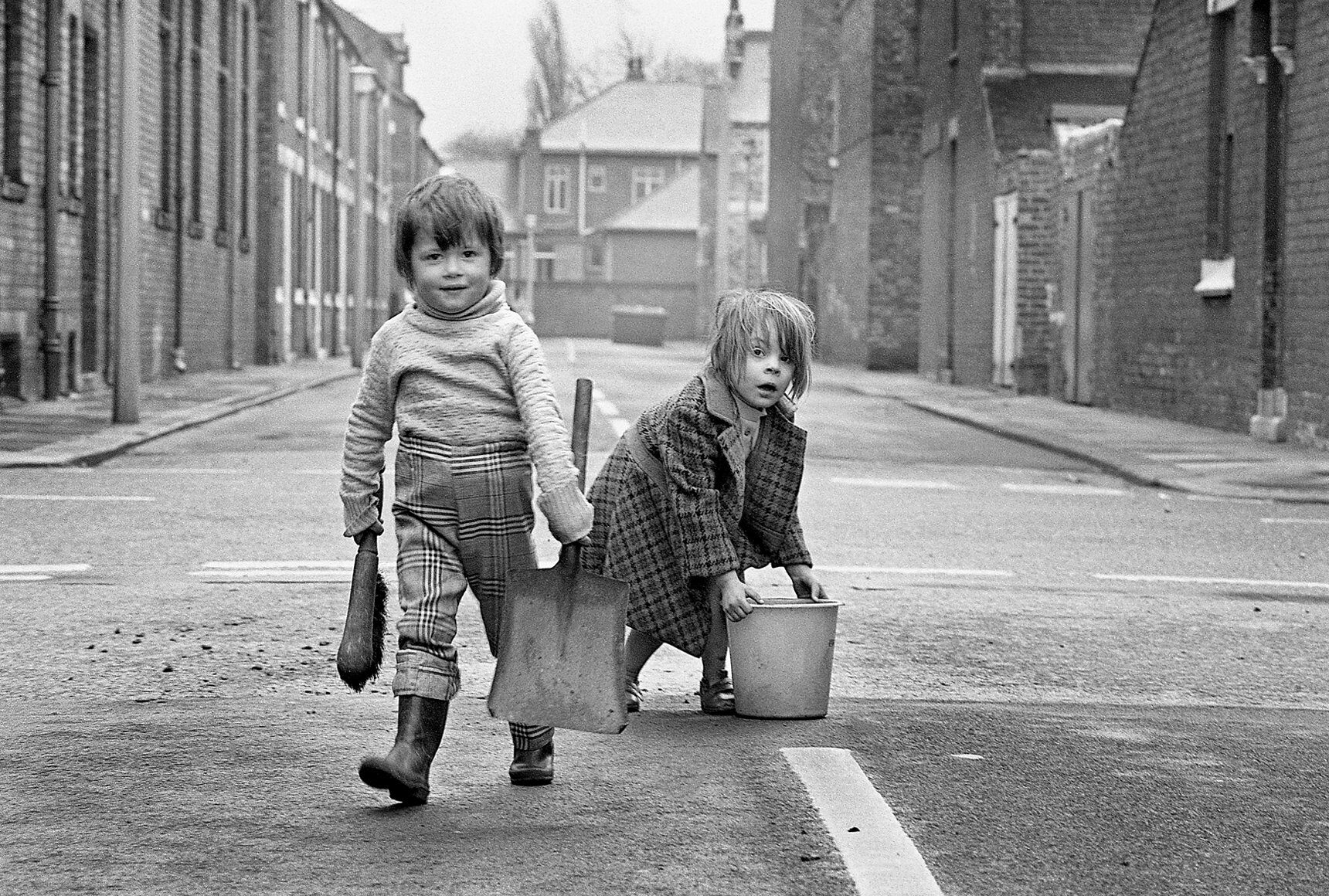 Two children carrying a bucket and a sweeping brush on a street in Teesside.